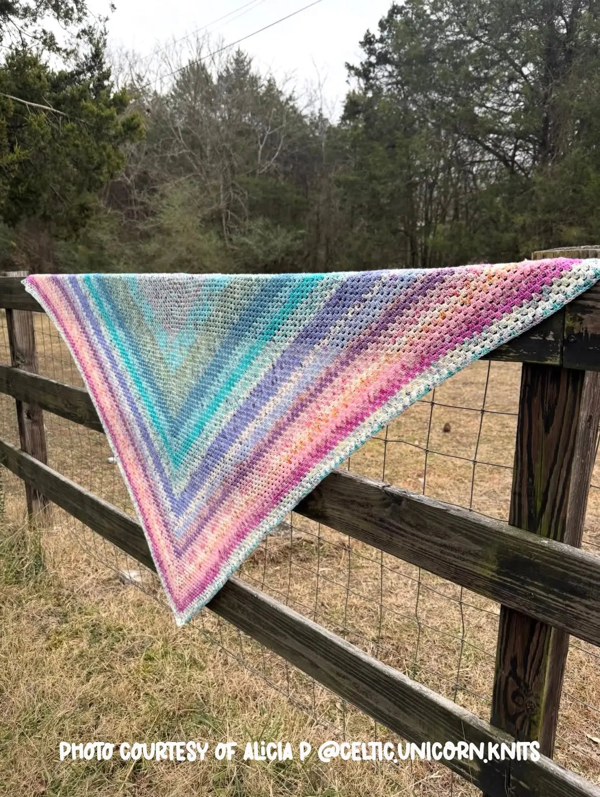 Crocheted shawl with colorful stripes draped over a wooden fence in a natural setting.