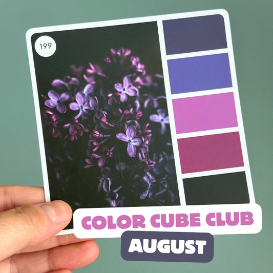 A hand holds a square printed card titled 'COLOR CUBE CLUB AUGUST' against a muted green background. The card features a photo of purple flowers on the left and five color swatches on the right, including shades of purple, blue-violet, pink, magenta, and black.