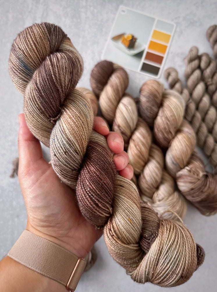 Hand holding skeins of brown and beige yarn with a color swatch in the background.